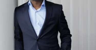 Man in a navy blazer and light blue open-collar shirt leans against a wall, one hand in his pocket, smiling slightly at the c
