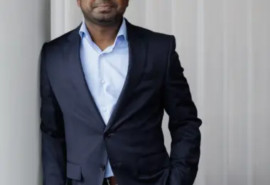Man in a navy blazer and light blue open-collar shirt leans against a wall, one hand in his pocket, smiling slightly at the c