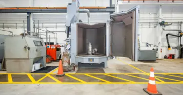 Industrial equipment inside a manufacturing facility, including an open grey testing chamber containing a submersible pump, with yellow floor markings and orange safety cones.