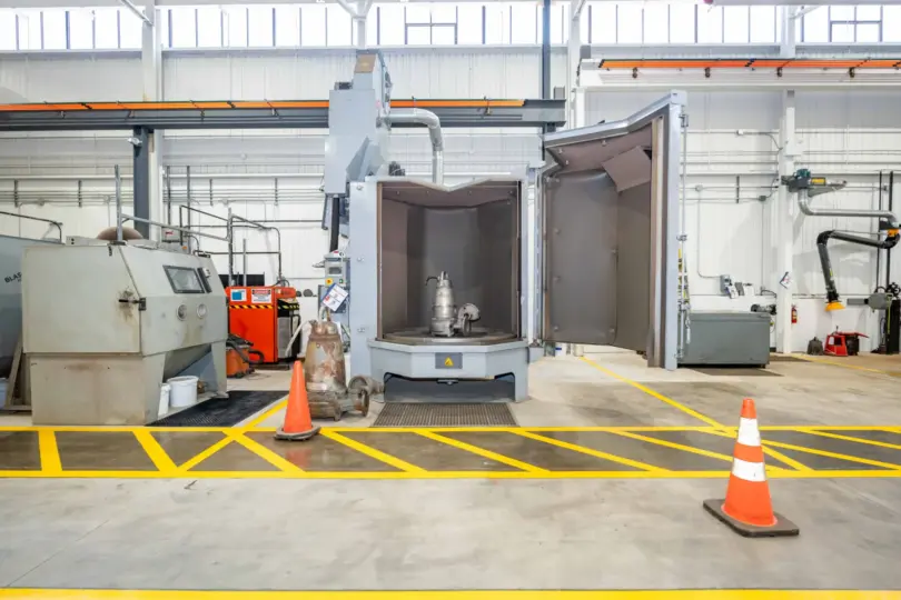 Industrial equipment inside a manufacturing facility, including an open grey testing chamber containing a submersible pump, with yellow floor markings and orange safety cones.