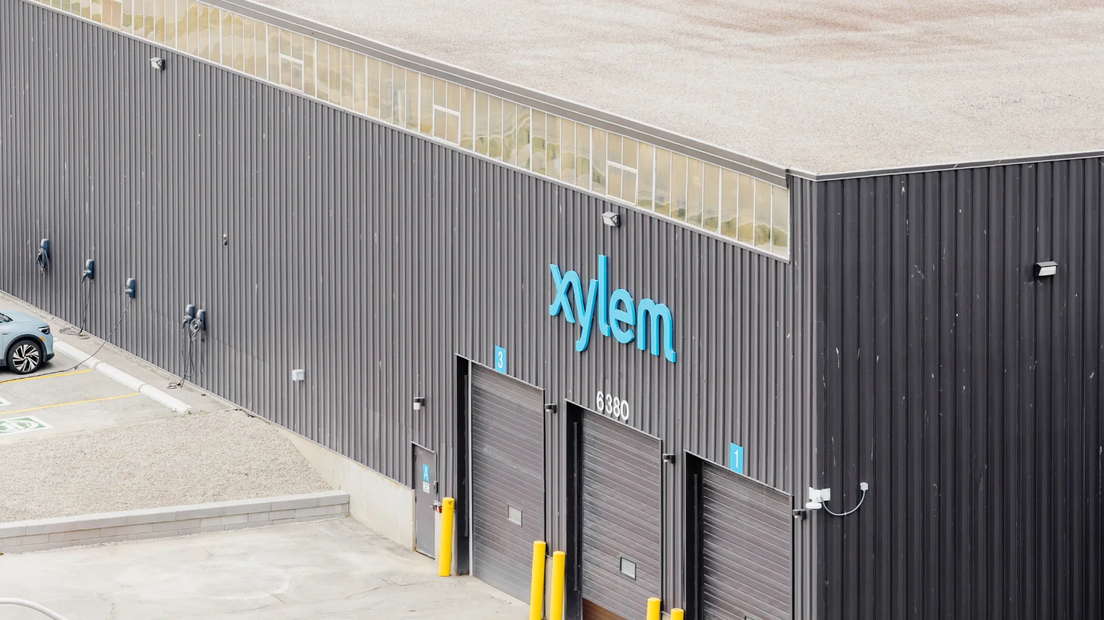 Xylem Toronto hub warehouse with blue Xylem logo, numbered loading bay doors, yellow bollards and EV chargers beside a parked